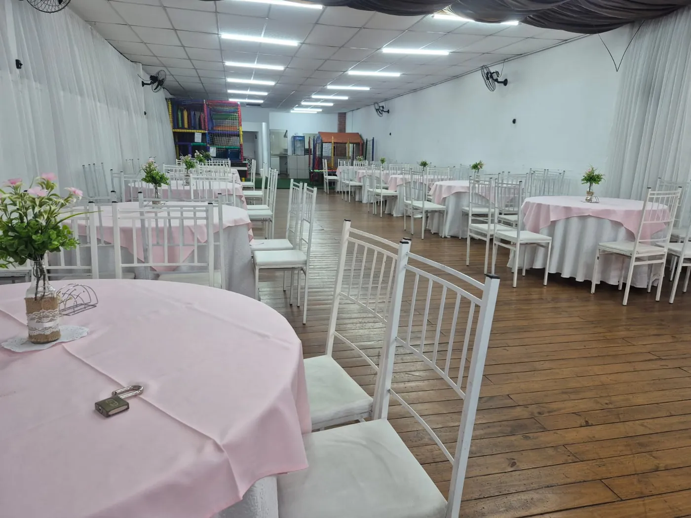 Salão com mesas de toalha rosa e cadeiras brancas prontas para a festa