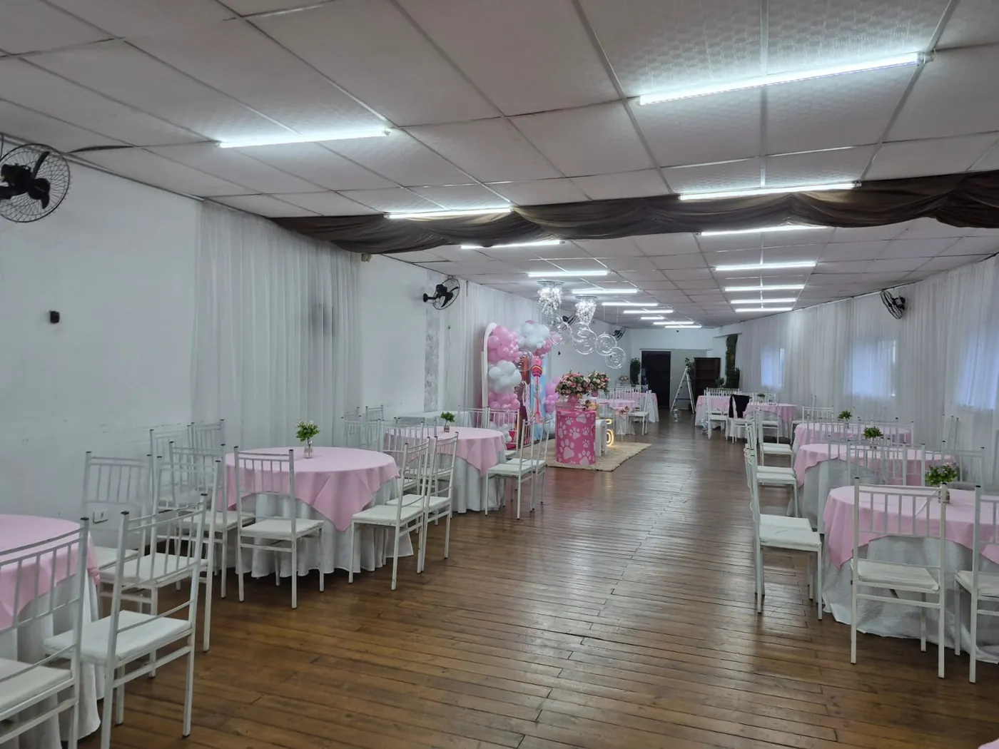 Salão com mesas de toalha rosa, cadeiras brancas e decoração de balões